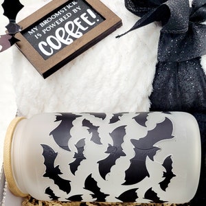Puede incluir: Una manta blanca y peluda con una bolsa de regalo negra y plateada atada con una cinta negra. Un peque&ntilde;o letrero de madera con el texto "My broomstick is powered by coffee!" est&aacute; encima de la manta. Una taza de vidrio esmerilado con un patr&oacute;n de murci&eacute;lagos negros est&aacute; en la esquina inferior derecha de la imagen.