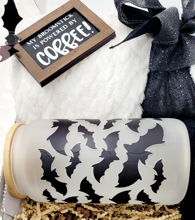 Puede incluir: Una manta blanca y peluda con una bolsa de regalo negra y plateada atada con una cinta negra. Un peque&ntilde;o letrero de madera con el texto "My broomstick is powered by coffee!" est&aacute; encima de la manta. Una taza de vidrio esmerilado con un patr&oacute;n de murci&eacute;lagos negros est&aacute; en la esquina inferior derecha de la imagen.