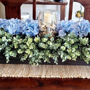 May include: A long wooden planter box filled with blue hydrangeas and green foliage. The planter is sitting on a brown woven table runner.