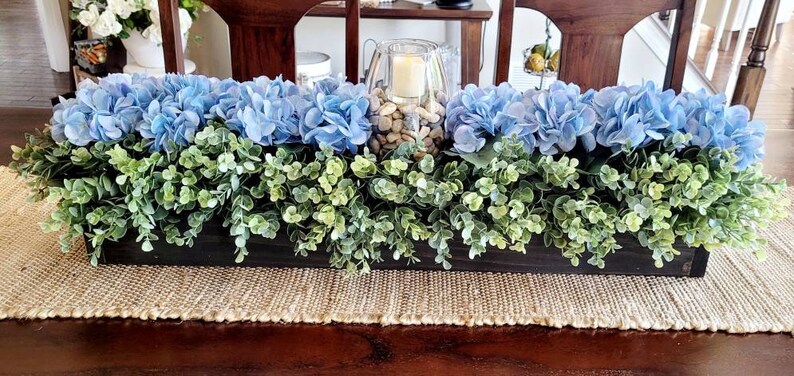 May include: A long wooden planter box filled with blue hydrangeas and green foliage. The planter is sitting on a brown woven table runner.