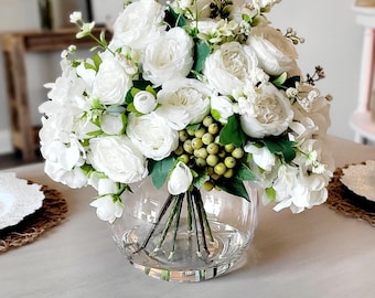 May include: A floral arrangement featuring white roses, hydrangeas, and small green berries in a clear, round glass vase. The bouquet includes green leaves and delicate white filler flowers, with the stems visible through the glass base.