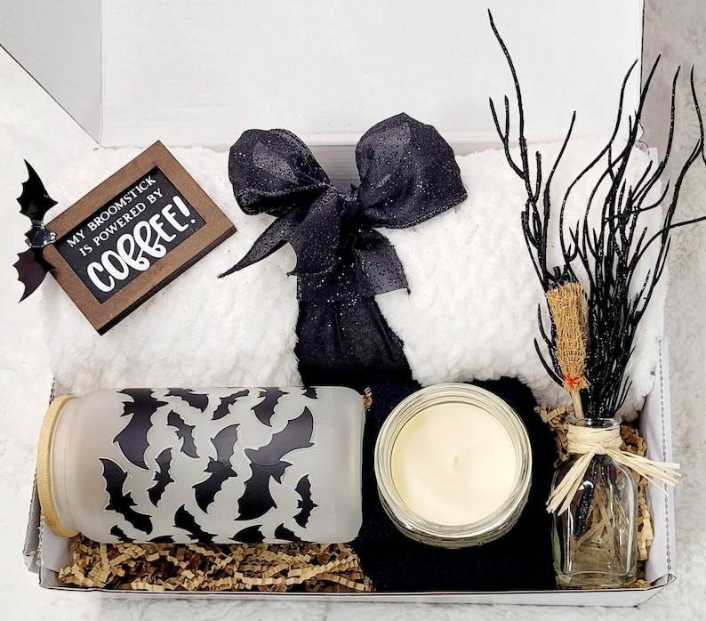 Op de afbeelding: Een cadeaudoos gevuld met een wit, pluche deken, een zwart-wit lint, een zwart-wit, mat glas met een vleermuispatroon, een doorzichtige glazen pot met een kaars en een doorzichtige glazen fles met een zwarte tak en een bruine bezem. De doos bevat ook een bruine kaart met de tekst "My broomstick is powered by coffee!"