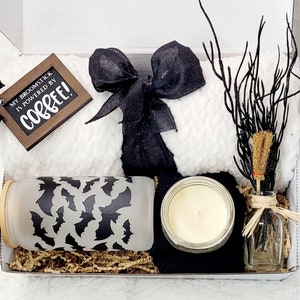 Puede incluir: Una caja de regalo blanca llena de una manta de lana con estampado blanco y negro, una cinta de sat&eacute;n negra, una vela de vidrio, un frasco de vidrio con un patr&oacute;n de murci&eacute;lago negro y una peque&ntilde;a botella de vidrio con una rama marr&oacute;n y un borla marr&oacute;n. La caja tambi&eacute;n incluye un letrero de madera que dice "My broomstick is powered by coffee!"