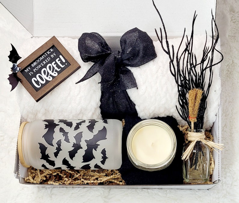 Puede incluir: Una caja de regalo blanca llena de una manta de lana con estampado blanco y negro, una cinta de sat&eacute;n negra, una vela de vidrio, un frasco de vidrio con un patr&oacute;n de murci&eacute;lago negro y una peque&ntilde;a botella de vidrio con una rama marr&oacute;n y un borla marr&oacute;n. La caja tambi&eacute;n incluye un letrero de madera que dice "My broomstick is powered by coffee!"