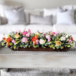 May include: A wooden trough filled with a floral arrangement of pink, orange, and white flowers. The flowers are surrounded by green foliage and are sitting on a white coffee table.