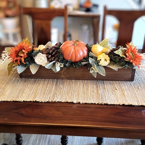 May include: A wooden box centerpiece with fall decor. The box is filled with greenery, white flowers, orange and yellow pumpkins, and sunflowers.
