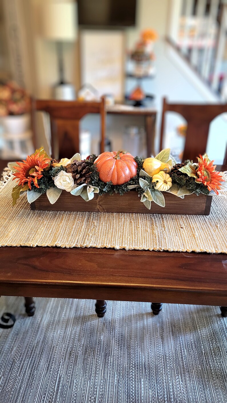 May include: A wooden box centerpiece with fall decor. The box is filled with greenery, white flowers, orange and yellow pumpkins, and sunflowers.
