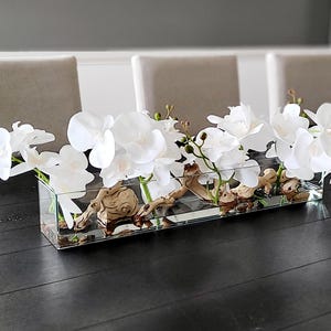 May include: A rectangular glass vase filled with white orchid flowers and driftwood. The vase sits on a dark wooden table. The flowers are in full bloom, and the driftwood adds a natural element to the arrangement.