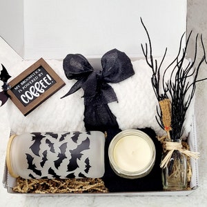 Puede incluir: Una caja de regalo blanca llena de una variedad de art&iacute;culos, que incluyen una manta blanca peluda con una cinta negra, un frasco de vidrio con un patr&oacute;n de murci&eacute;lago negro, una vela en un frasco de vidrio y una peque&ntilde;a botella de vidrio con una rama seca dentro. La caja tambi&eacute;n incluye un letrero de madera que dice "My broomstick is powered by coffee!"