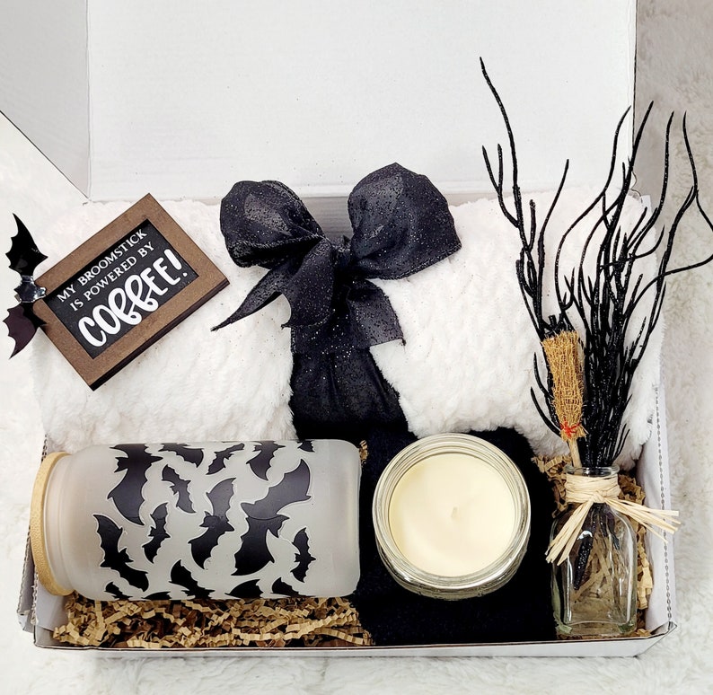 Puede incluir: Una caja de regalo blanca llena de una variedad de art&iacute;culos, que incluyen una manta blanca peluda con una cinta negra, un frasco de vidrio con un patr&oacute;n de murci&eacute;lago negro, una vela en un frasco de vidrio y una peque&ntilde;a botella de vidrio con una rama seca dentro. La caja tambi&eacute;n incluye un letrero de madera que dice "My broomstick is powered by coffee!"