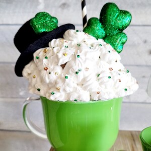 May include: A green ceramic mug with a white whipped cream topping, decorated with green shamrock sprinkles and a black top hat with a green shamrock. The mug has a clear glass handle and a black and white striped straw.