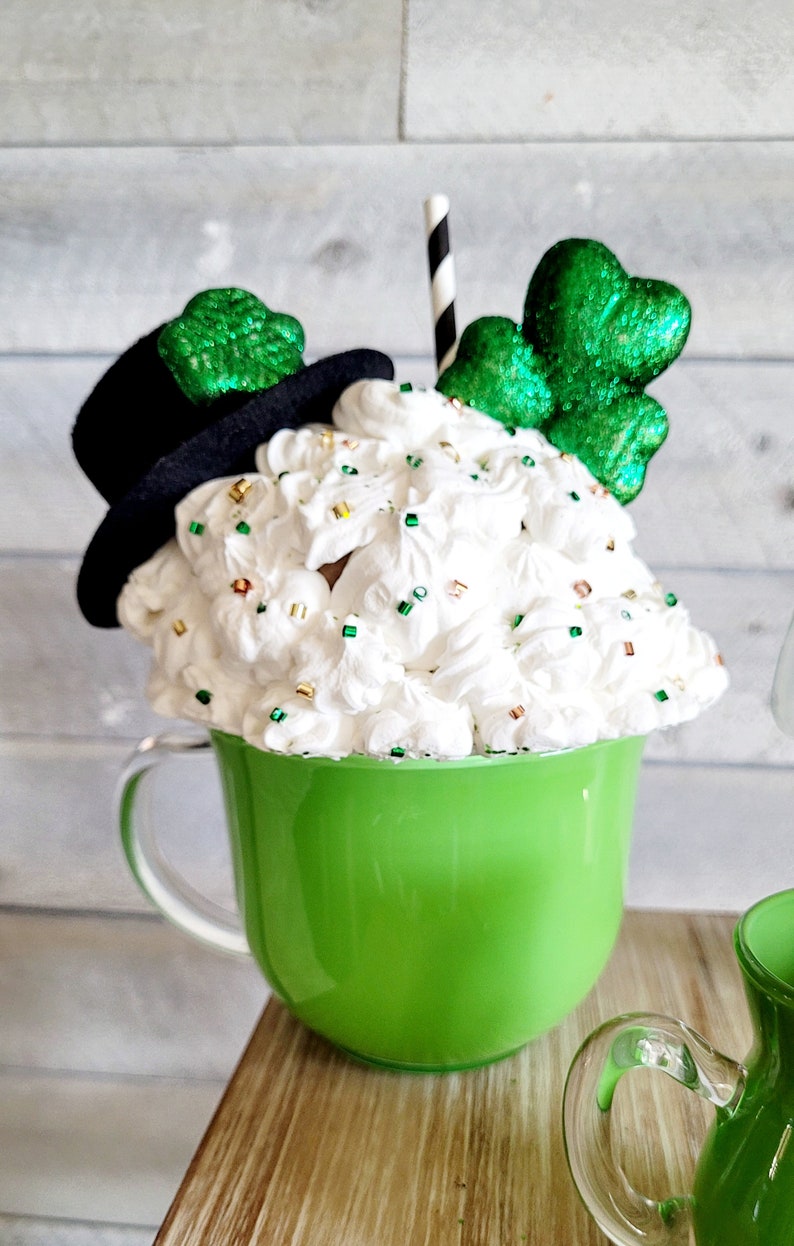 May include: A green ceramic mug with a white whipped cream topping, decorated with green shamrock sprinkles and a black top hat with a green shamrock. The mug has a clear glass handle and a black and white striped straw.