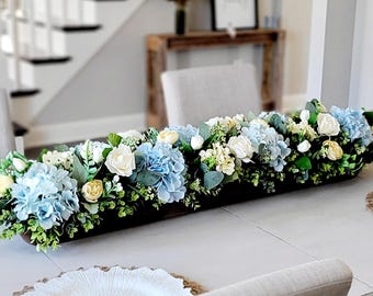 May include: A long, dark wooden dough bowl centerpiece filled with an arrangement of light blue hydrangeas, cream-colored roses, small yellow flower buds, and various green eucalyptus leaves and foliage, creating a dense floral display.