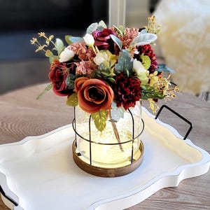 May include: A clear glass jar with a black wire cage and a wooden base, illuminated by internal warm string lights. The jar contains an arrangement of artificial flowers in shades of deep red, burnt orange, cream, and green foliage.