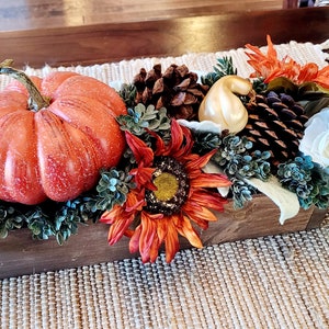 May include: A wooden box centerpiece with a variety of fall decor, including a large orange pumpkin, small white and gold gourds, pine cones, a sunflower, and white flowers.