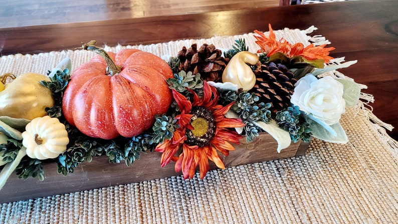 May include: A wooden box centerpiece with a variety of fall decor, including a large orange pumpkin, small white and gold gourds, pine cones, a sunflower, and white flowers.