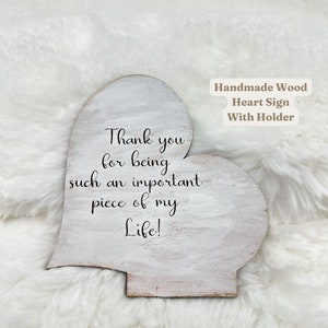 May include: A handmade wooden heart-shaped sign with a holder. The sign is painted white with distressed edges and features the text "Thank you for being such an important piece of my Life!" in black script.