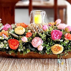 Puede incluir: Centro de mesa de madera lleno de flores artificiales de colores, incluyendo flores rosas, naranjas, amarillas y moradas. Un portavelas de cristal con una vela amarilla encendida se encuentra en el centro, rodeado de pequeñas piedras.