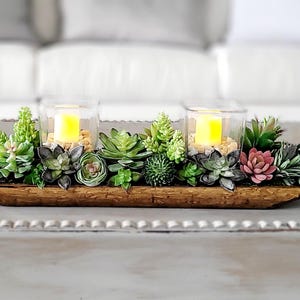 May include: A wooden centerpiece with two glass candle holders, each containing a yellow candle. The centerpiece is decorated with a variety of artificial succulents in shades of green and pink. The wooden base is approximately 18 inches long.