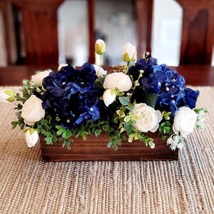 Op de afbeelding: Een rechthoekige houten kist gevuld met kunstbloemen. Het arrangement bevat diepblauwe hortensia's, witte rozen en groene bladeren. De kist staat op een beige geweven placemat.