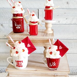 May include: Four Valentine's Day themed mugs with whipped cream and red hearts. The mugs are on two wooden tiered stands. The mugs say "kisses 25c", "love in a mug", and "love".