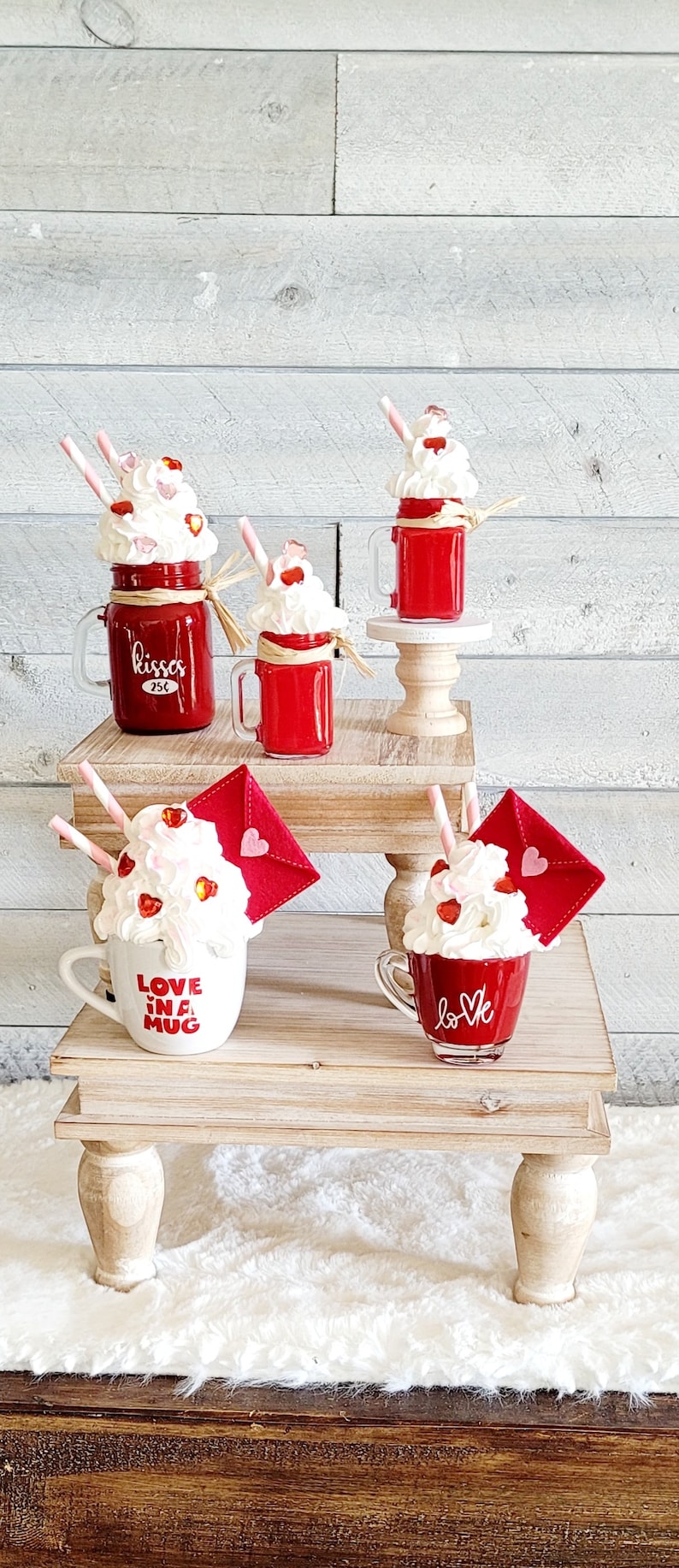May include: Four Valentine's Day themed mugs with whipped cream and red hearts. The mugs are on two wooden tiered stands. The mugs say "kisses 25c", "love in a mug", and "love".