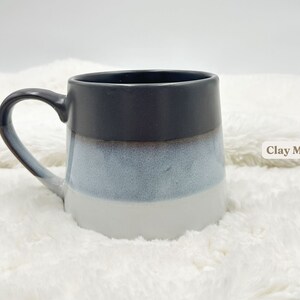 May include: A grey, white and blue ceramic mug with a rounded shape and a handle. The mug is sitting on a white fluffy surface. "Clay Mug" is written in brown text on the right side of the image.