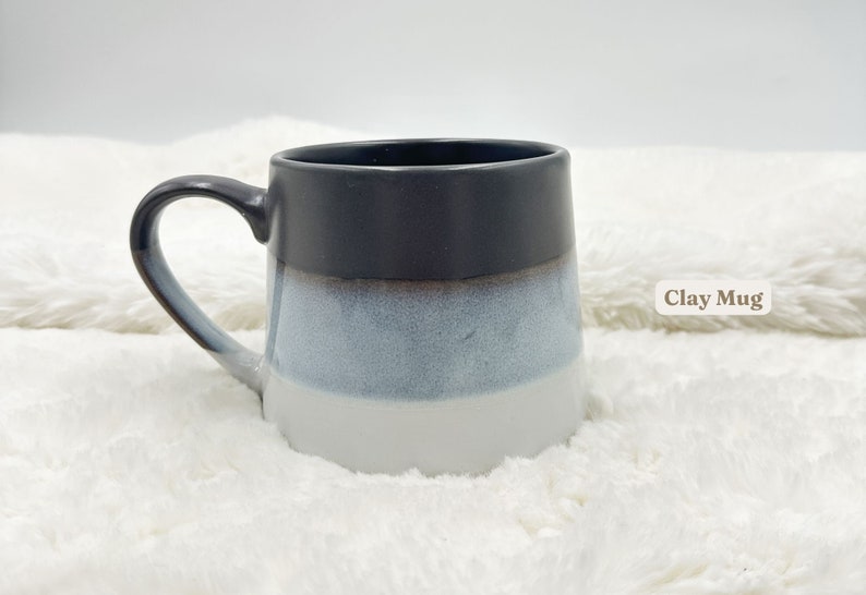 May include: A grey, white and blue ceramic mug with a rounded shape and a handle. The mug is sitting on a white fluffy surface. "Clay Mug" is written in brown text on the right side of the image.