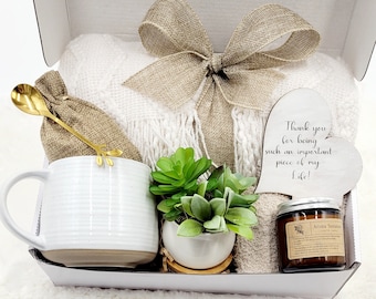 May include: A gift box filled with a white blanket, a white mug, a gold spoon, a small succulent plant, a candle, and a heart-shaped wooden sign. The sign reads, "Thank you for being such an important piece of my Life!" The box is tied with a burlap bow.