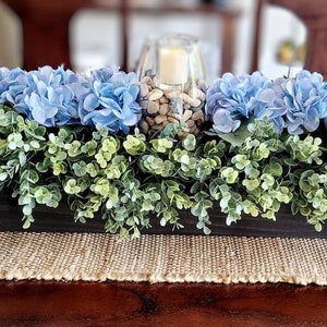 May include: A wooden box centerpiece with blue hydrangea flowers and green foliage. The centerpiece is decorated with a glass vase filled with decorative stones.