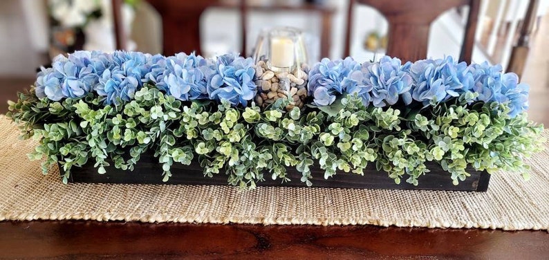 May include: A wooden box centerpiece with blue hydrangea flowers and green foliage. The centerpiece is decorated with a glass vase filled with decorative stones.