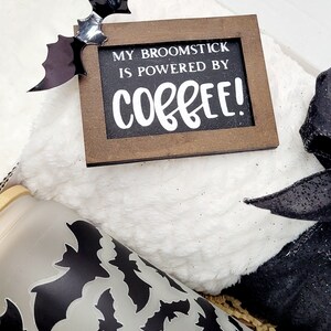 Puede incluir: Una caja de regalo con tem&aacute;tica de Halloween con una manta blanca peluda, una cinta negra y blanca, un vaso transparente con un patr&oacute;n de murci&eacute;lago negro y un letrero de madera que dice "My broomstick is powered by coffee!"