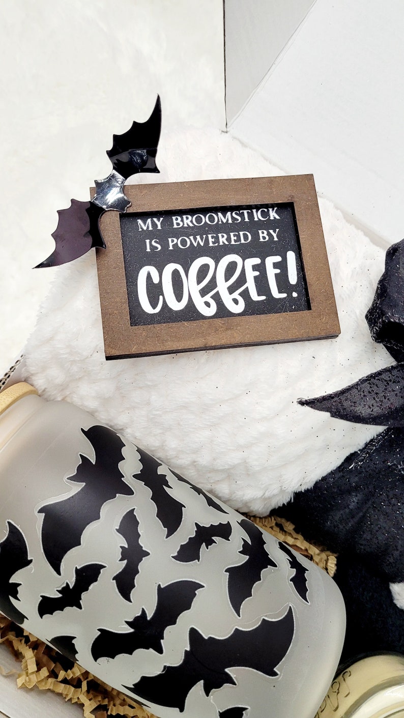Puede incluir: Una caja de regalo con tem&aacute;tica de Halloween con una manta blanca peluda, una cinta negra y blanca, un vaso transparente con un patr&oacute;n de murci&eacute;lago negro y un letrero de madera que dice "My broomstick is powered by coffee!"