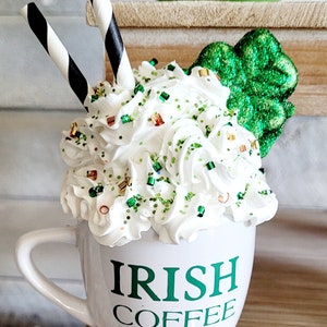 May include: White ceramic mug with green lettering that reads "Irish Coffee". The mug is filled with whipped cream and green and gold sprinkles. Two black and white striped straws are sticking out of the whipped cream. A green shamrock is on the side of the mug.