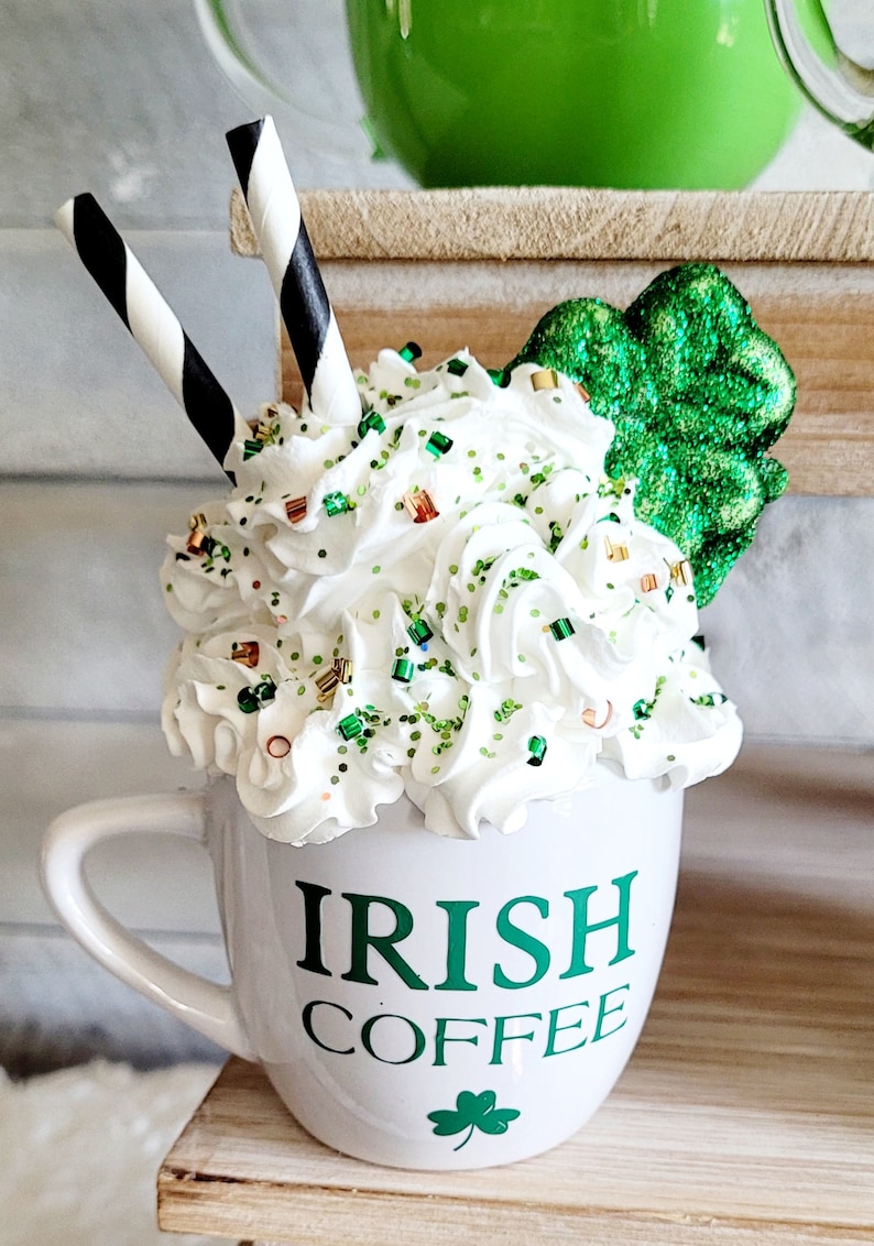 May include: White ceramic mug with green lettering that reads "Irish Coffee". The mug is filled with whipped cream and green and gold sprinkles. Two black and white striped straws are sticking out of the whipped cream. A green shamrock is on the side of the mug.