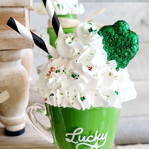 May include: A green mug with white whipped cream, green sprinkles, and a green shamrock on top. The mug has the word "Lucky" written on it in white.