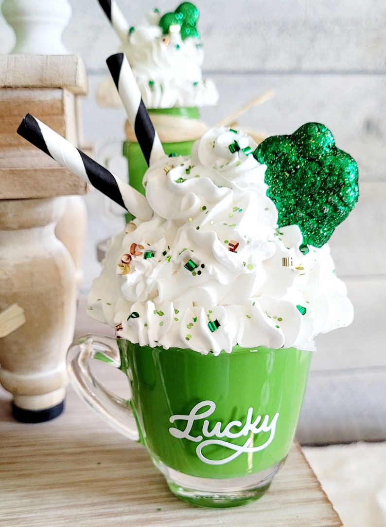 May include: A green mug with white whipped cream, green sprinkles, and a green shamrock on top. The mug has the word "Lucky" written on it in white.