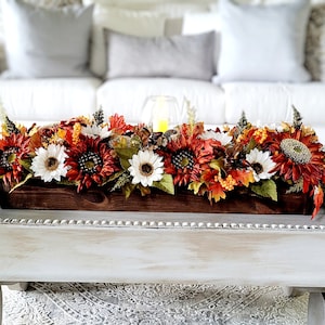 May include: A wooden tray with a white distressed finish and a beaded trim sits on a rug. The tray holds a wooden box filled with a fall floral arrangement of sunflowers, white daisies, and orange and red leaves.