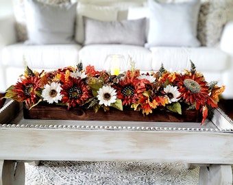 May include: A wooden tray with a white distressed finish and a beaded trim sits on a rug. The tray holds a wooden box filled with a fall floral arrangement of sunflowers, white daisies, and orange and red leaves.