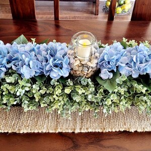 May include: A blue hydrangea centerpiece with greenery and a glass candle holder filled with white stones. The centerpiece is on a burlap runner.