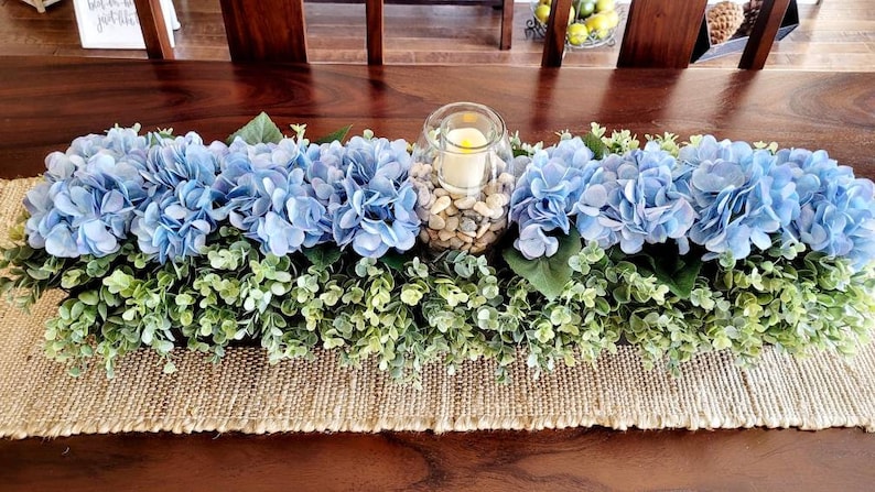 May include: A blue hydrangea centerpiece with greenery and a glass candle holder filled with white stones. The centerpiece is on a burlap runner.