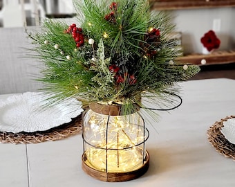 May include: A clear glass jar lantern with a dark wood base and top, encased in a black wire frame. The jar is filled with warm white fairy lights and topped with a festive arrangement of green pine needles, red berries, and small white beads.