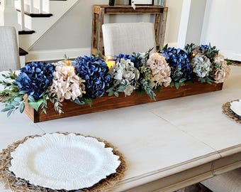 May include: A long, rectangular dark brown wooden box centerpiece filled with a dense arrangement of deep blue and light beige hydrangea blossoms, green foliage, and small glass candle holders containing glowing yellow tea lights.