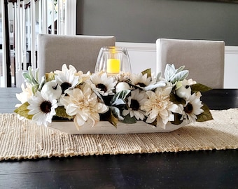 May include: A white wooden centerpiece filled with cream-colored sunflowers, dahlias, and greenery. A glass candle holder with a yellow candle sits in the center. The arrangement rests on a jute table runner.