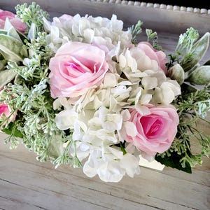 May include: A white wooden box filled with a floral arrangement of white hydrangeas and pink roses. The flowers are surrounded by green foliage.