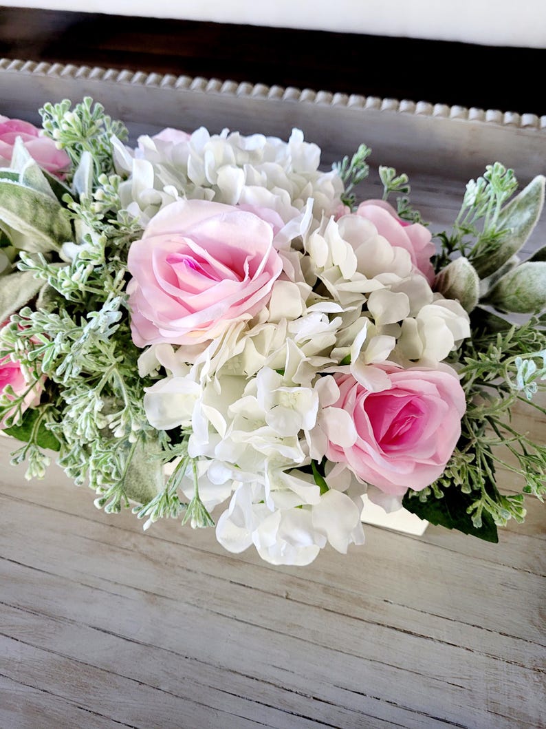 May include: A white wooden box filled with a floral arrangement of white hydrangeas and pink roses. The flowers are surrounded by green foliage.