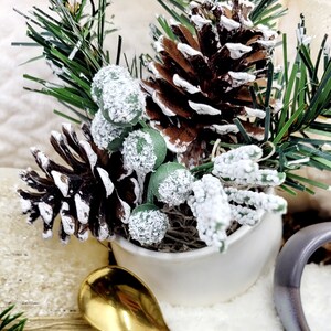 May include: A white ceramic pot filled with a snowy pine sprig, pine cones, and green berries. The pot is sitting on a white surface with a gold spoon next to it.