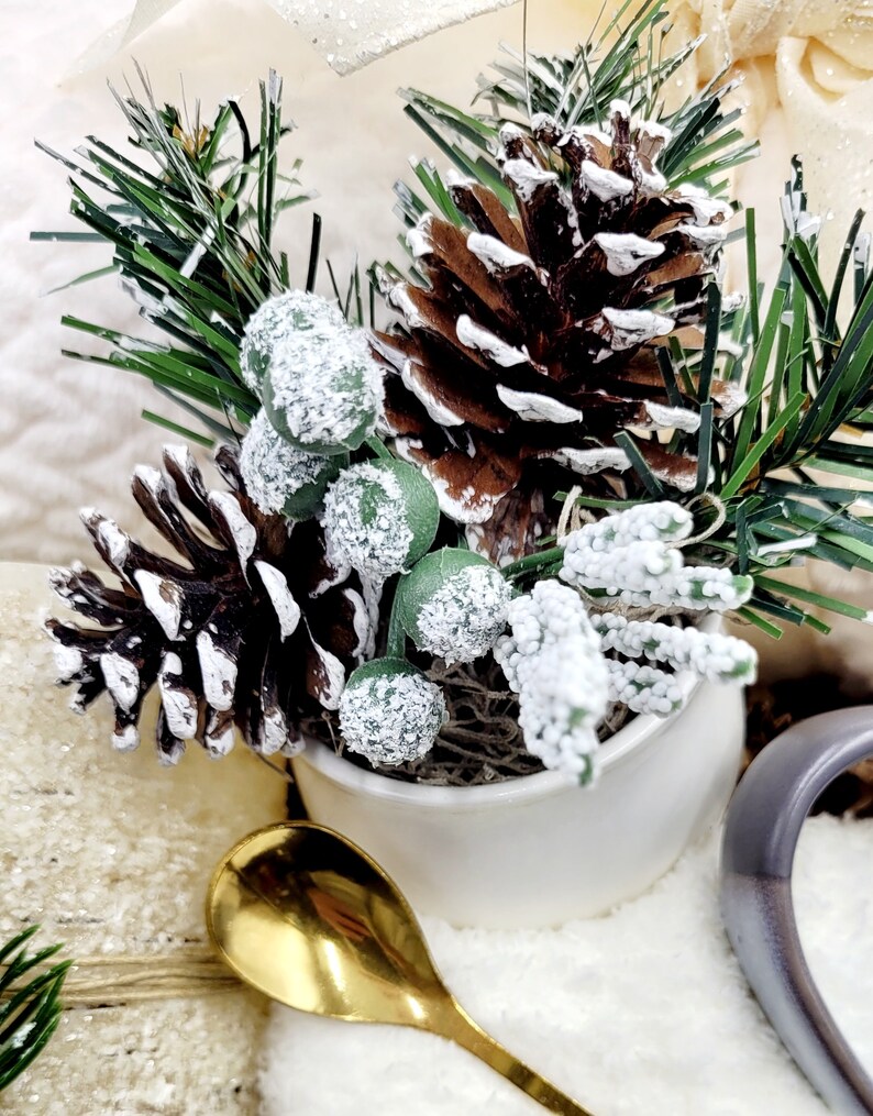 May include: A white ceramic pot filled with a snowy pine sprig, pine cones, and green berries. The pot is sitting on a white surface with a gold spoon next to it.