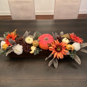 May include: A wooden box centerpiece with fall-themed decor. It features a bright orange pumpkin, white and cream pumpkins, pine cones, sunflowers, and fall-colored leaves.
