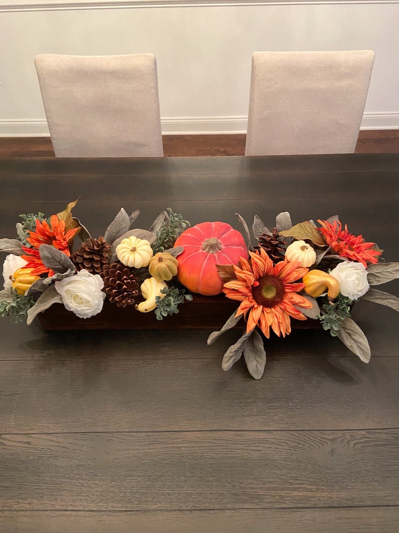 May include: A wooden box centerpiece with fall-themed decor. It features a bright orange pumpkin, white and cream pumpkins, pine cones, sunflowers, and fall-colored leaves.